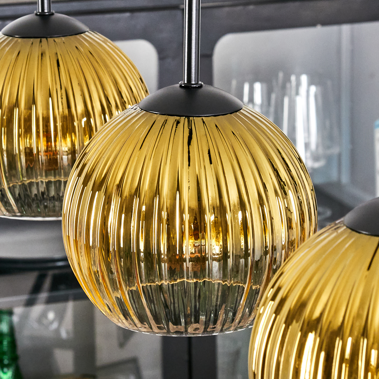 Drei goldene, gerippte Glas-Pendelleuchten hängen an schwarzen Stangen. Der Hintergrund zeigt einen verschwommenen Schrank mit Gläsern und grünen Flaschen.