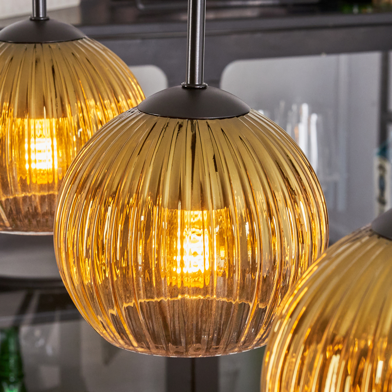 Drei goldfarbene, gerippte Glas-Pendelleuchten sind beleuchtet. Die Leuchten sind an dunklen Metallvorrichtungen aufgehängt, mit unscharfem Hintergrund.