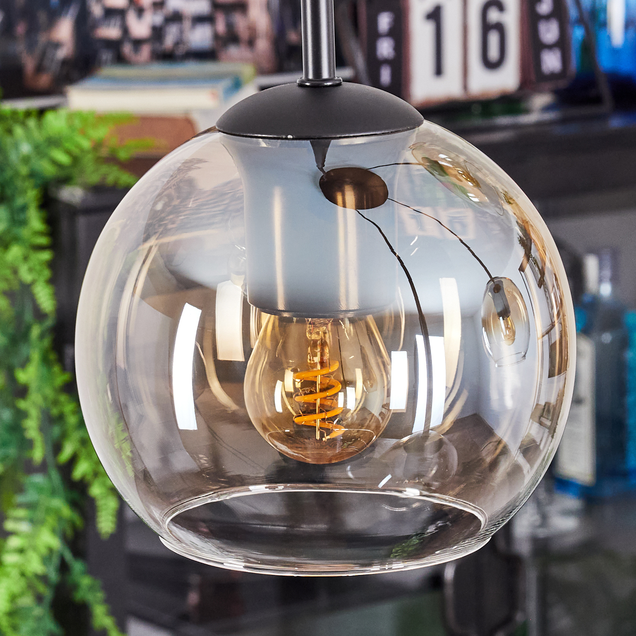 Eine moderne Glashängelampe mit einer leuchtenden Glühbirne. Der Hintergrund zeigt einen Kalender und Pflanzen, was auf eine Innenraumumgebung hindeutet.