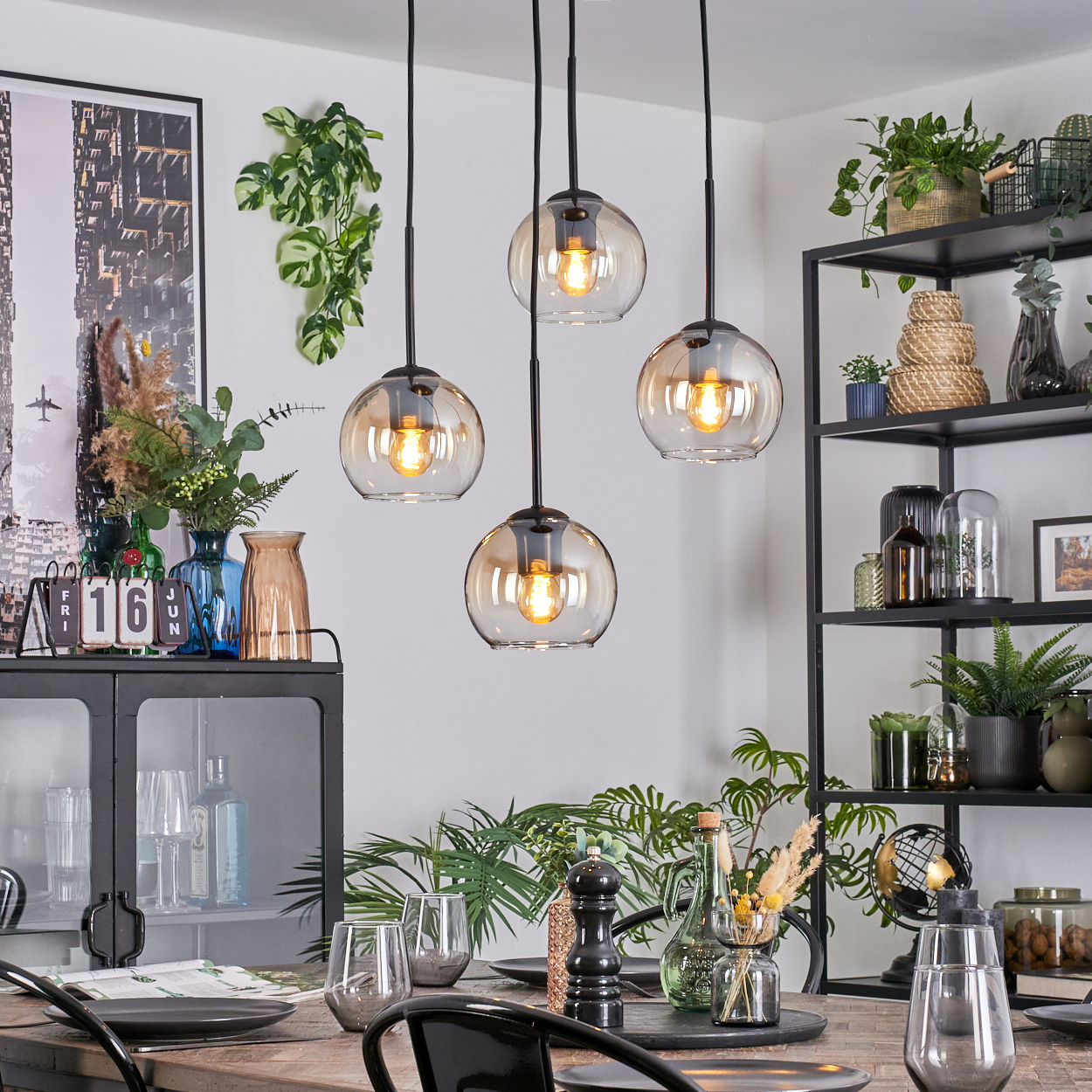 Esszimmer mit Hängelampen. Tisch mit Gläsern und Tellern gedeckt. Regale mit Pflanzen und Dekoration. Vasen und Bilder an den Wänden.
