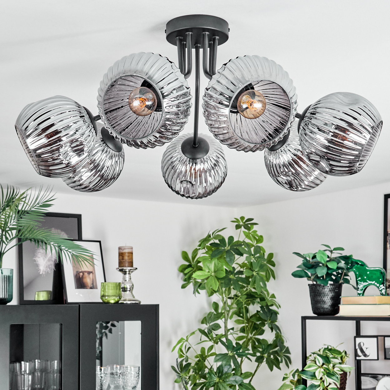 Eine moderne Deckenlampe mit fünf silbernen Glasschirmen. Darunter ein schwarzer Schrank, Pflanzen und Dekorationen.
