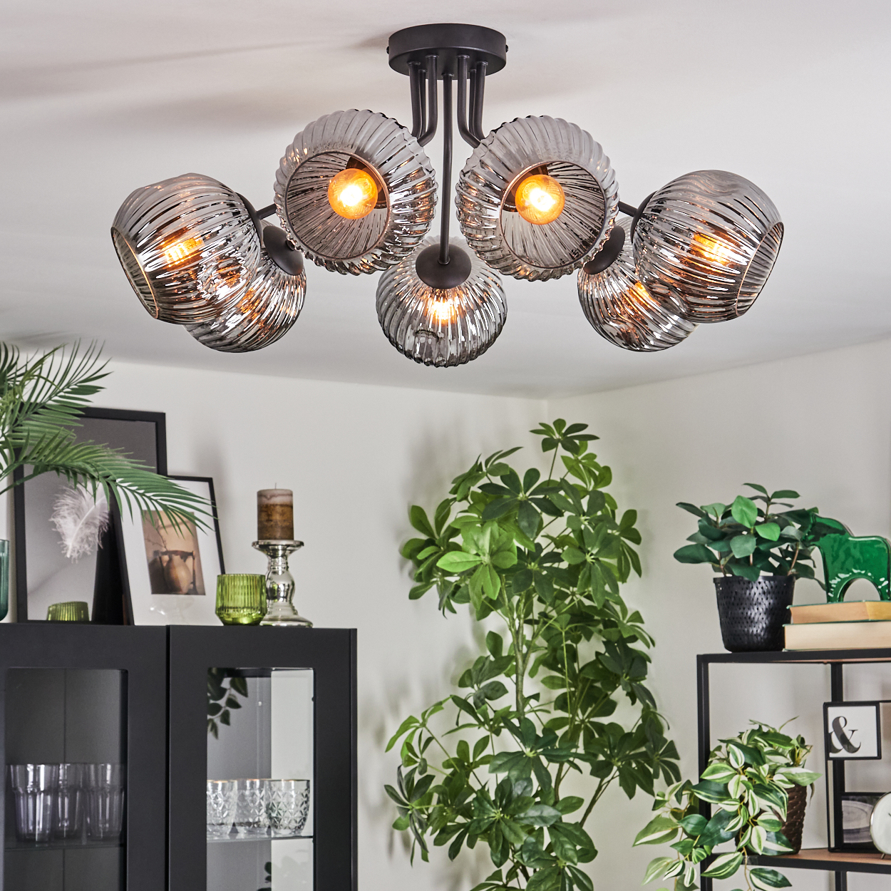 Eine moderne Deckenleuchte mit sechs kugelförmigen Lichtern. Der Raum hat Pflanzen, einen schwarzen Schrank und ein Regal. Die Decke ist weiß.