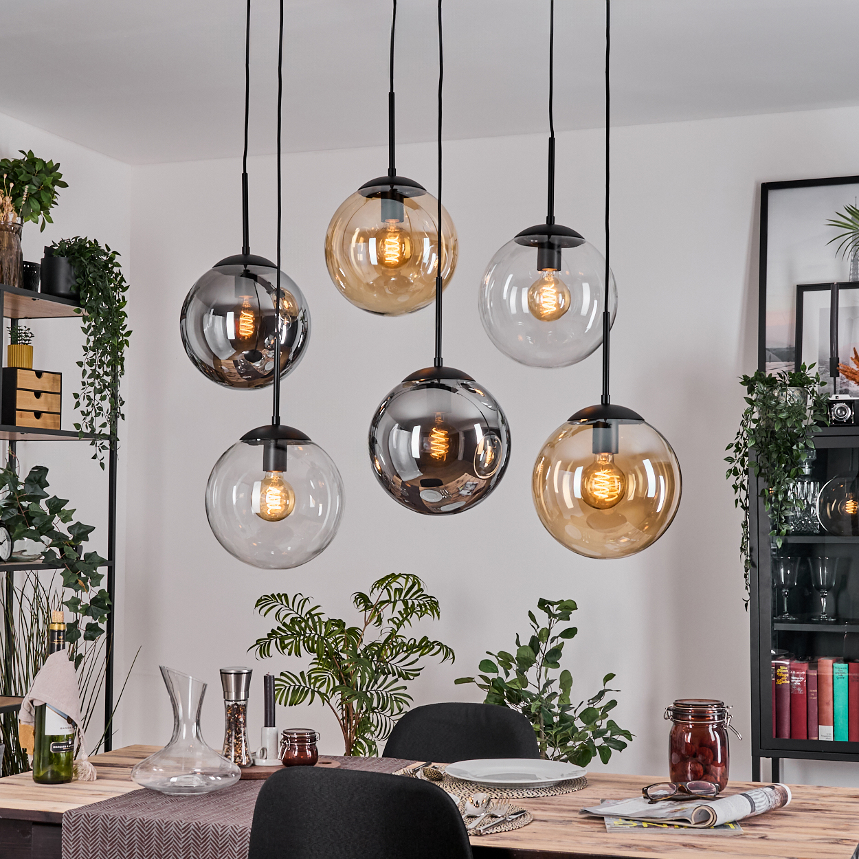 Esszimmer mit sechs runden Pendelleuchten. Ein Tisch ist mit Tellern, Gläsern und einem Glas gesetzt. Pflanzen und Dekoration sind sichtbar.