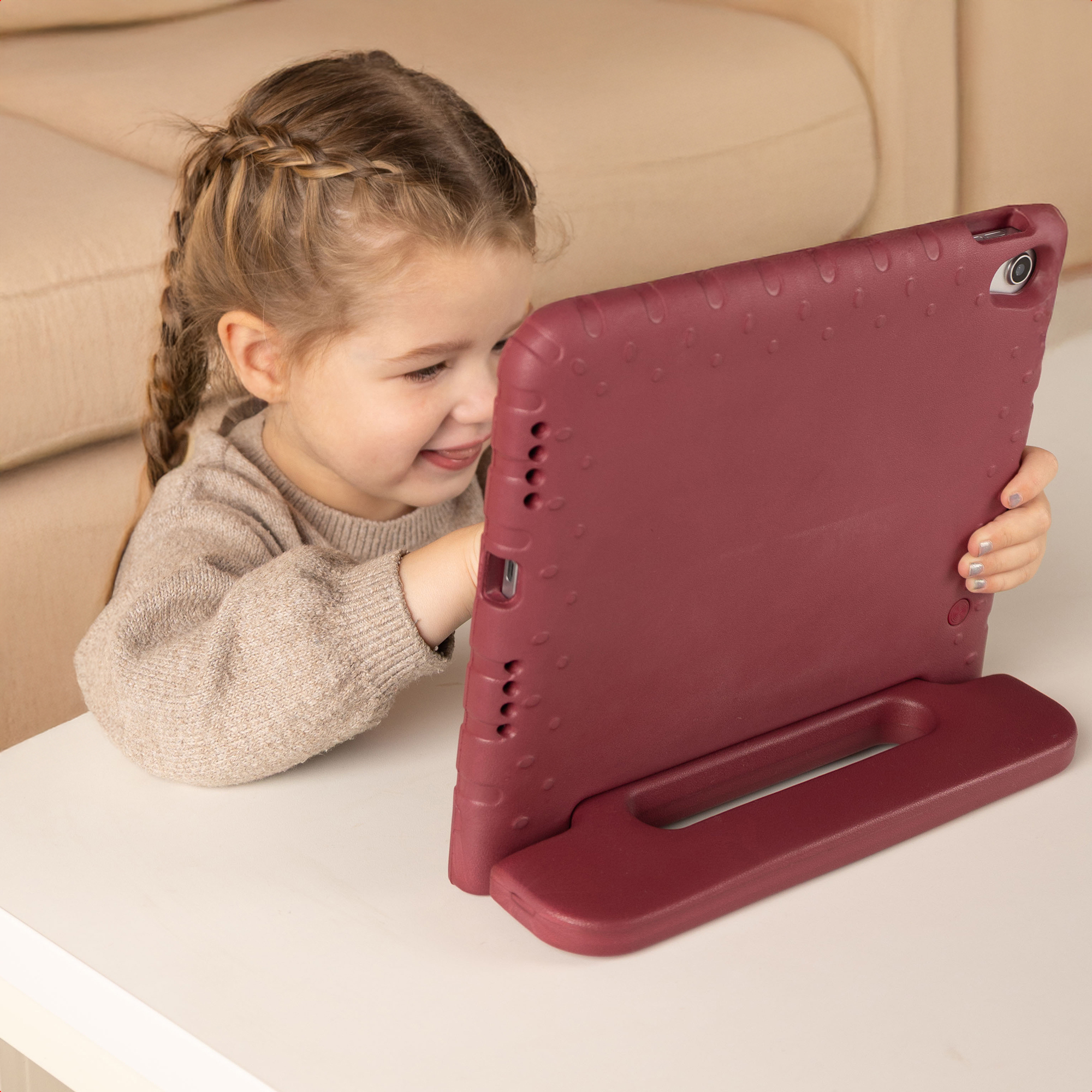 Een jong meisje met vlechten glimlacht terwijl ze naar een tablet in een rode hoes kijkt, zittend aan een witte tafel.