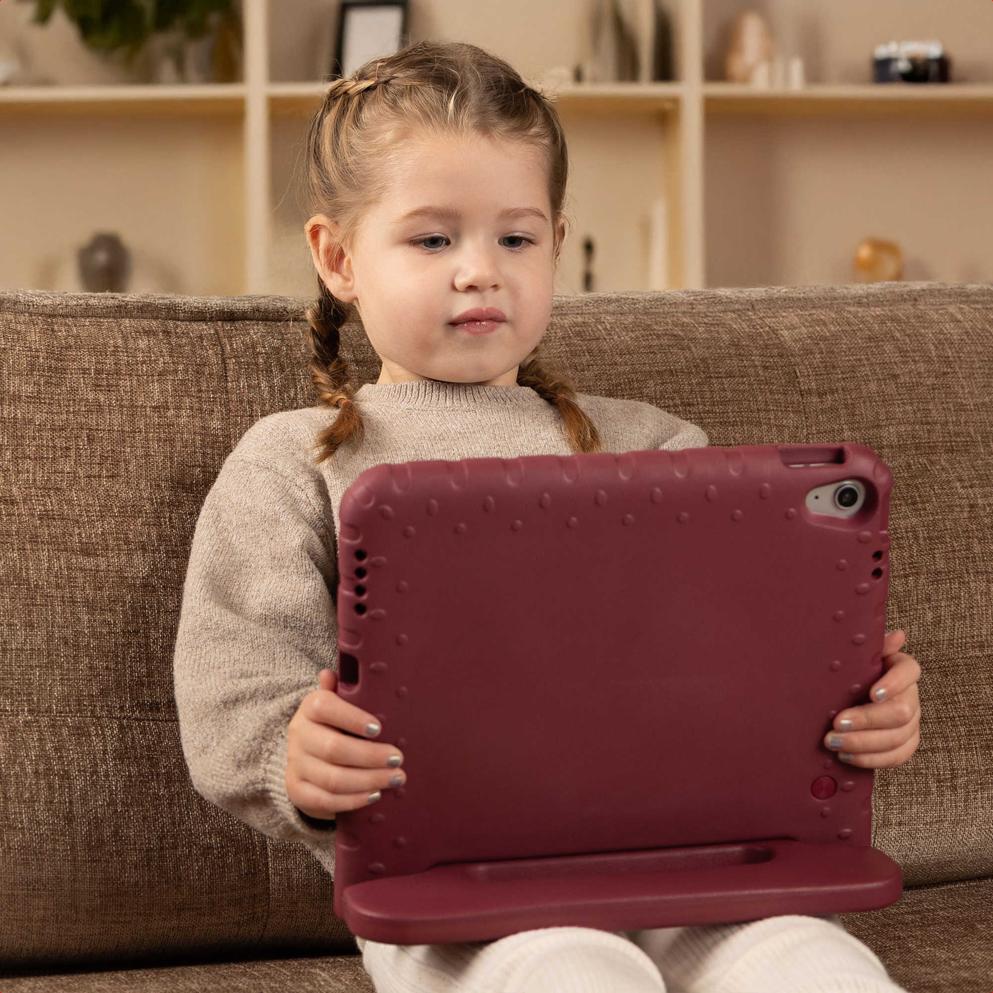 Een jong meisje met vlechten zit op een bank en houdt een bordeauxrode tablet vast. Ze draagt een trui. Achtergrond: planken.
