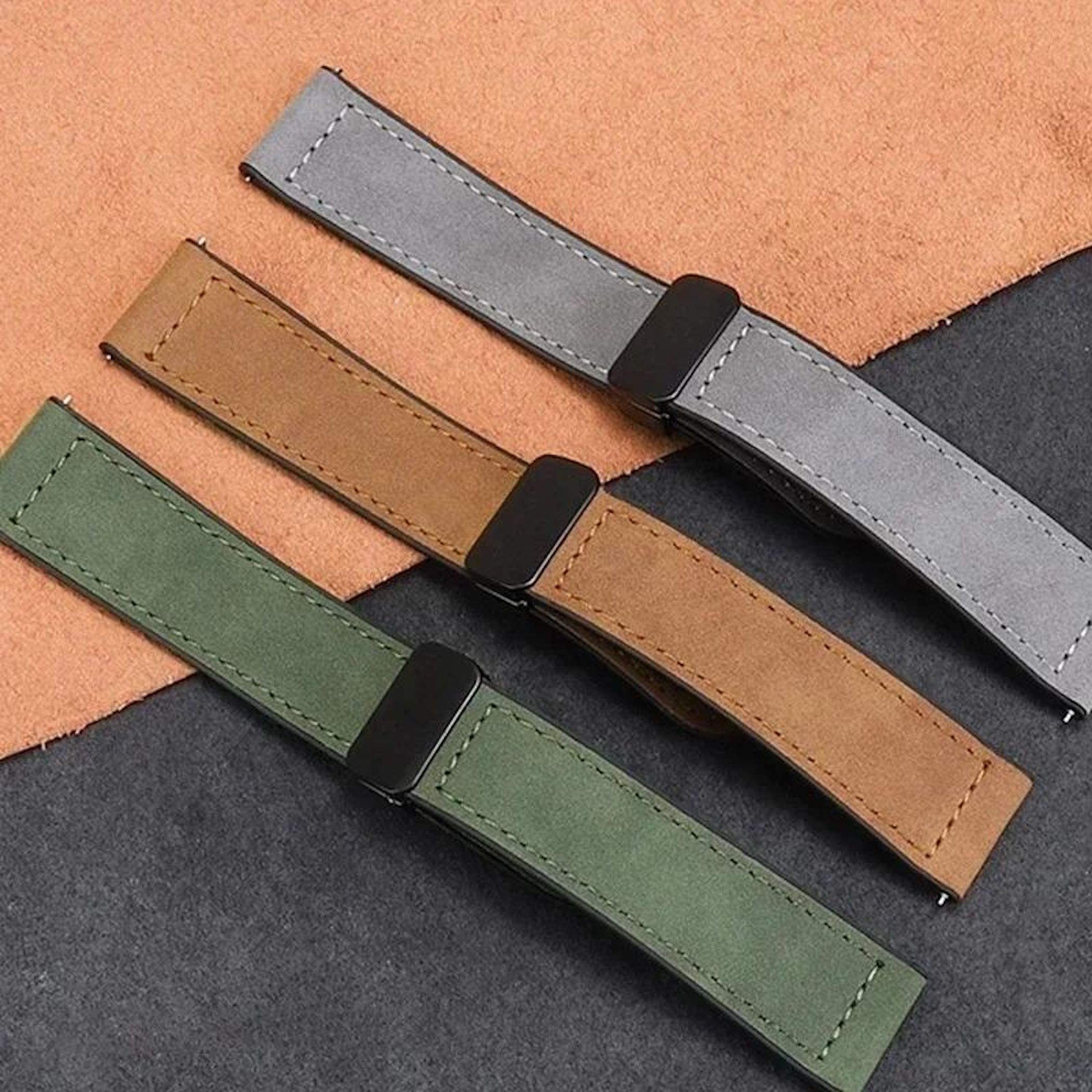Drei Leder-Uhrenarmbänder: grau, braun und grün. Sie liegen auf Oberflächen unterschiedlicher Farben. Schwarze Verschlüsse sind sichtbar.
