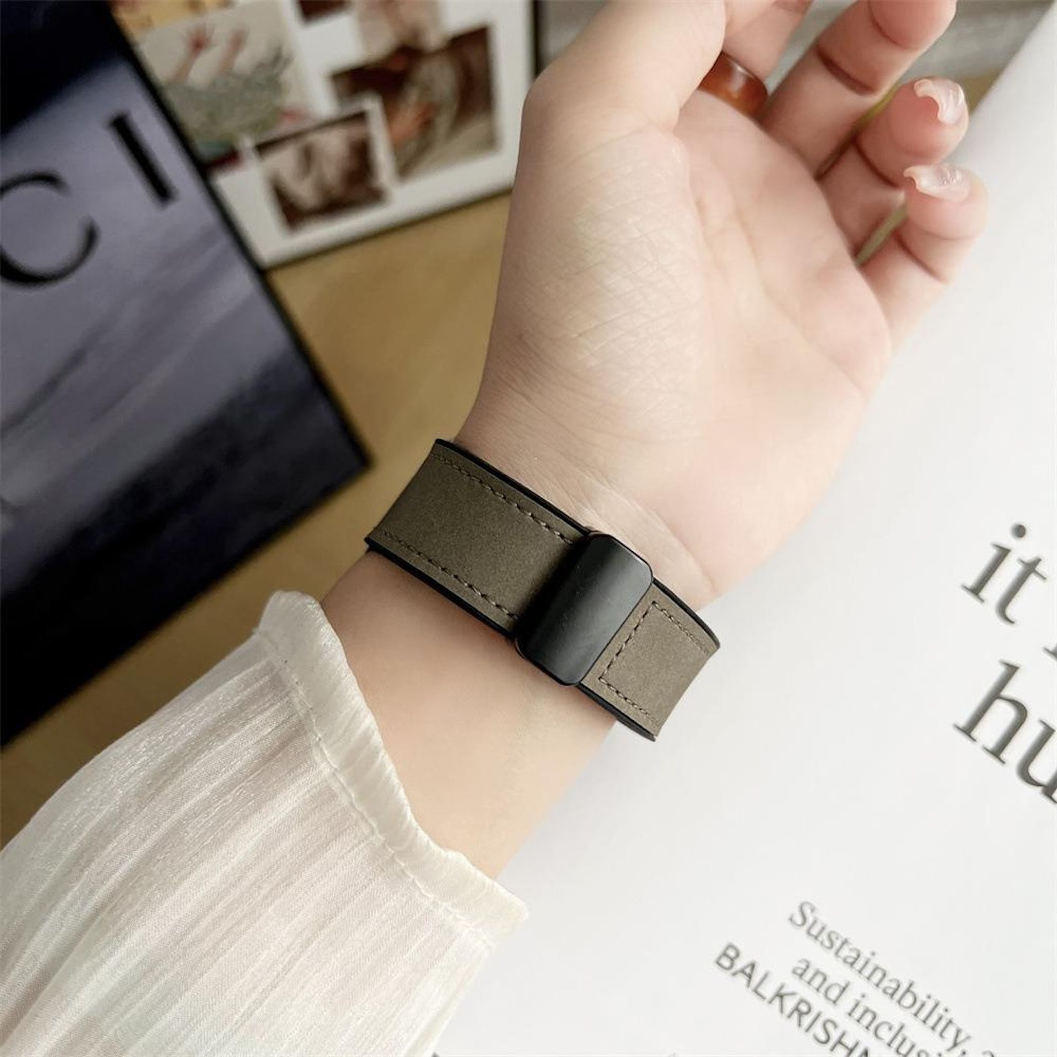 Ein Handgelenk mit grauem Armband wird gezeigt. Ein weißer Ärmel ist sichtbar. Text und Bilder sind im Hintergrund.