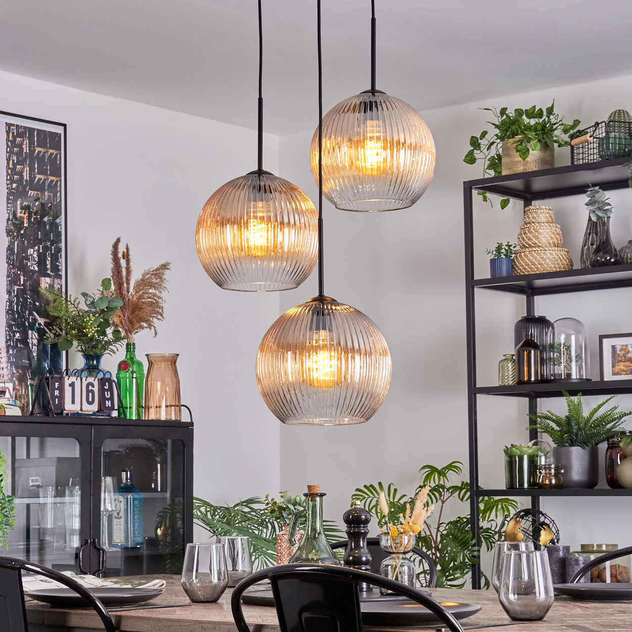 Esszimmer mit drei Hängelampen. Tisch mit Gläsern gedeckt. Regale mit Pflanzen und Dekoration im Hintergrund.