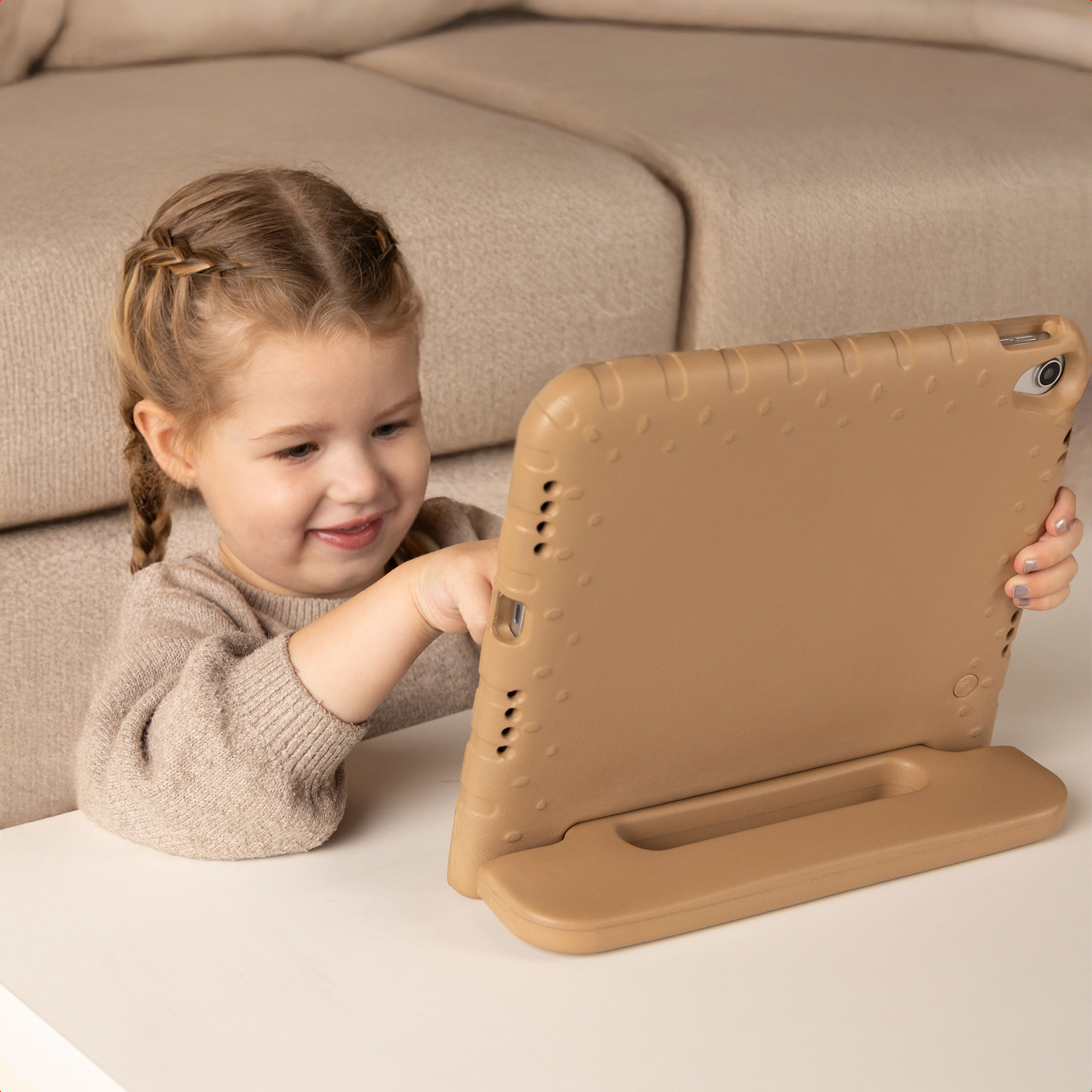 A smiling child with braids uses a tablet with a tan case. Background is a beige sofa and white table.