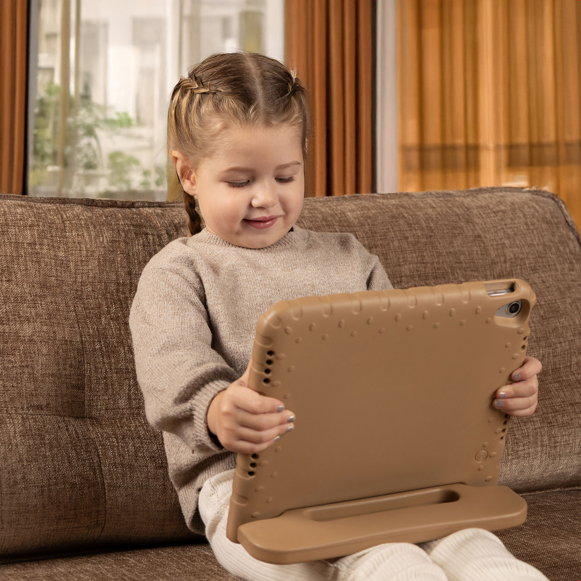 A girl with braids sits on a couch, holding a tablet. She looks at the screen. The background has curtains and a window.