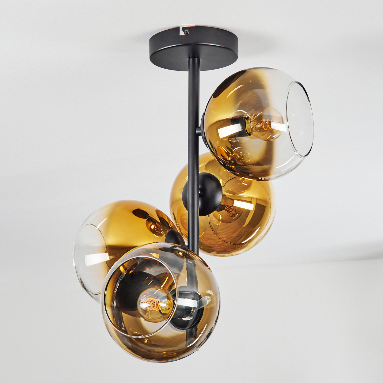 Eine moderne Deckenlampe mit vier bernsteinfarbenen Glaskugeln, in denen sich jeweils eine Glühbirne befindet. Die Leuchte ist schwarz, vor einem weißen Hintergrund.