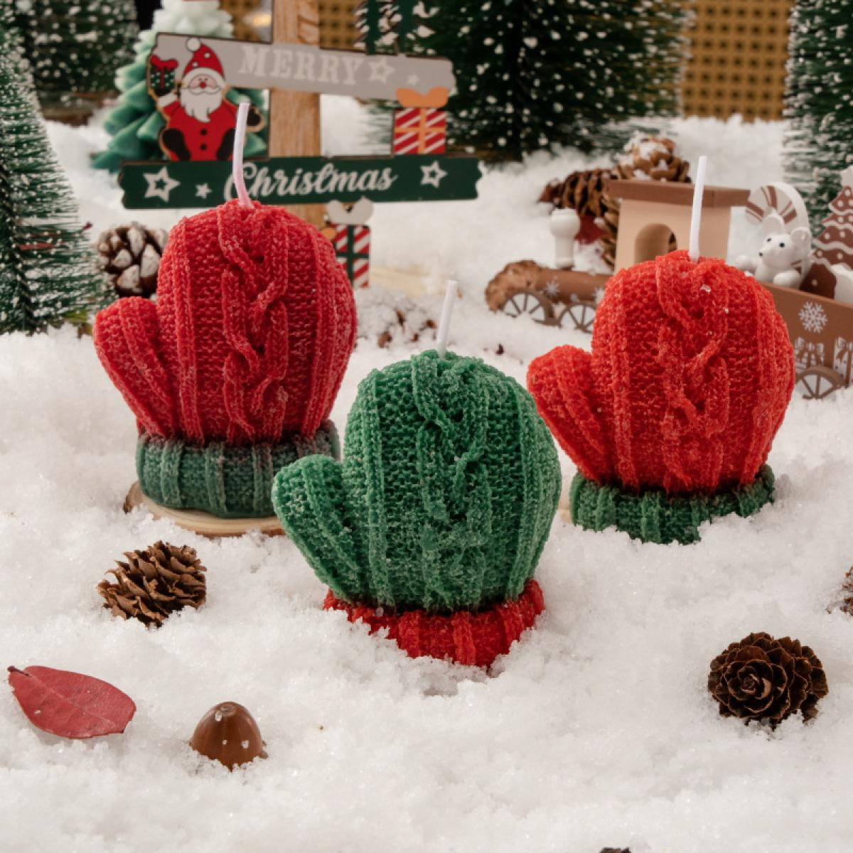 Drei handschuhförmige Kerzen in Rot und Grün auf einer schneebedeckten Oberfläche, mit einem Weihnachtsschild im Hintergrund.
