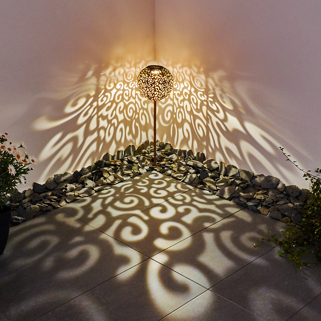 Eine dekorative Gartenlampe wirft Muster auf Boden und Wand. Umgeben von Steinen und Pflanzen, mit warmem Licht.