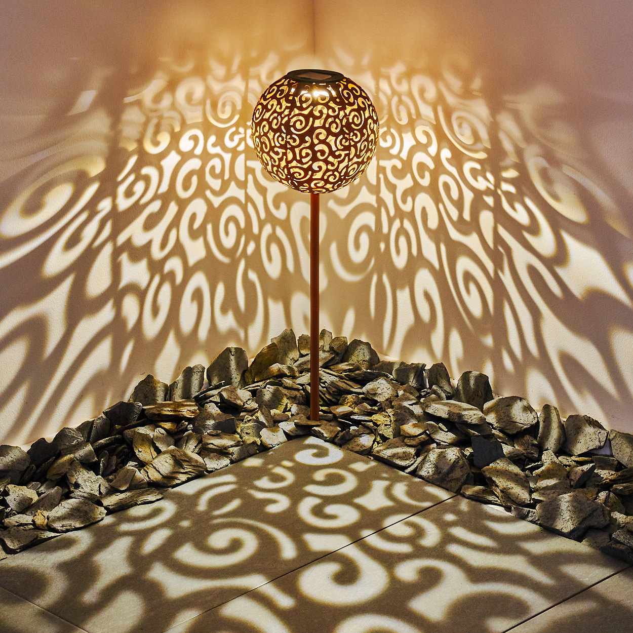 Eine dekorative Lampe mit einem gemusterten Globus wirft wirbelnde Schatten auf Wände und Boden. Es ist von Steinen umgeben.