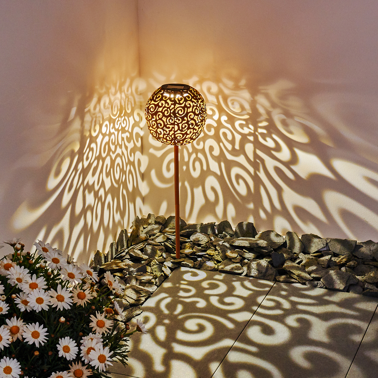 Eine dekorative Gartenlampe wirft Schattenmuster auf eine Wand. Gänseblümchen sind im Vordergrund. Ein Steinrand ist in der Mitte.