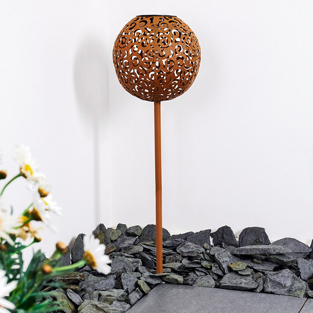 Eine dekorative Gartenleuchte mit einem gemusterten Ball auf einem Ständer, umgeben von Steinen und Blumen, vor einer weißen Wand.