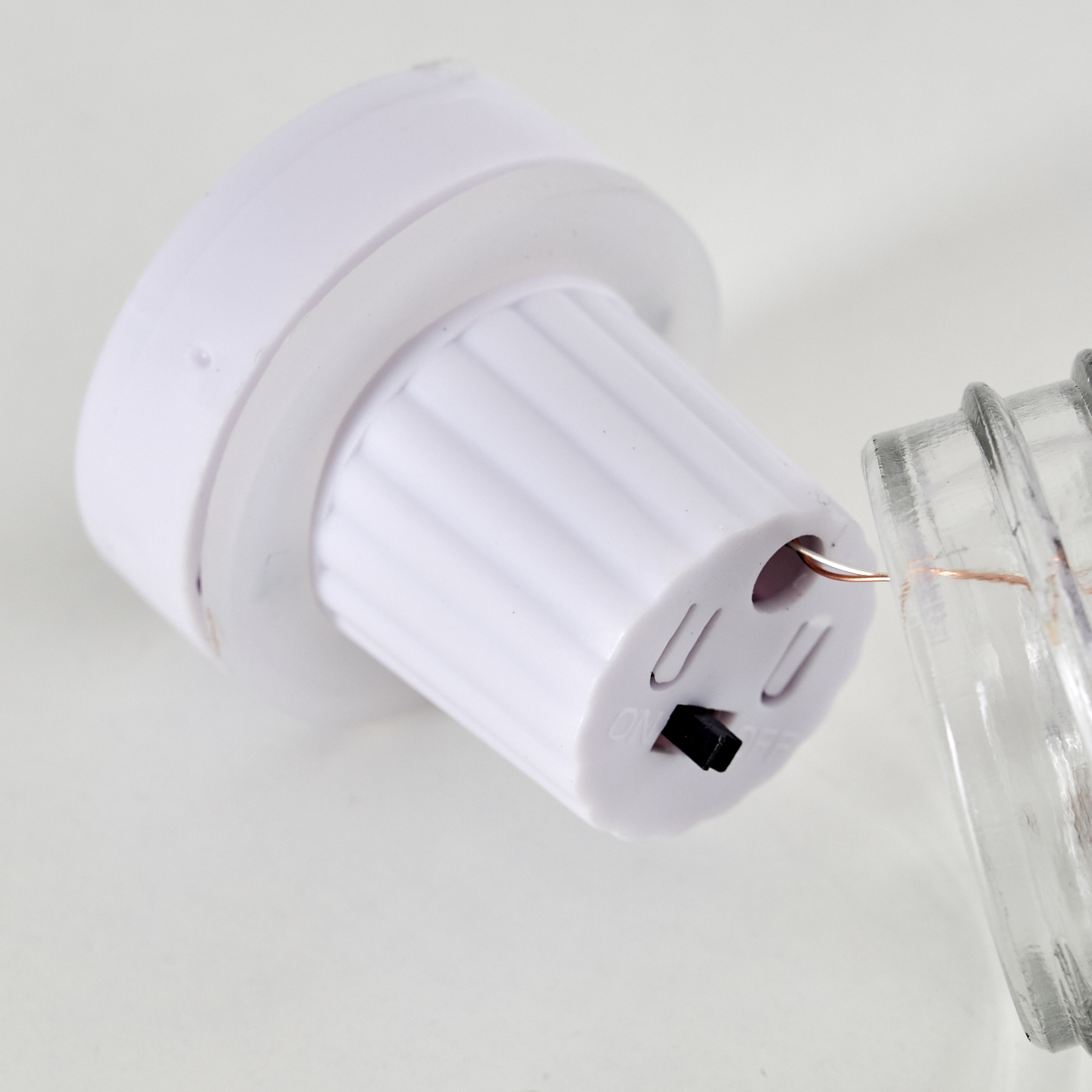 Weiße batteriebetriebene Lichtkomponente und ein Glas auf weißer Oberfläche. Die Lichtkomponente hat einen Ein/Aus-Schalter.