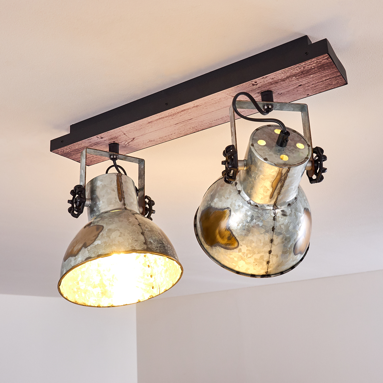 Zwei metallische Strahler, die an einem Holzbalken befestigt sind und an einer weißen Decke montiert sind. Die Lichter sind an und beleuchten den Raum.