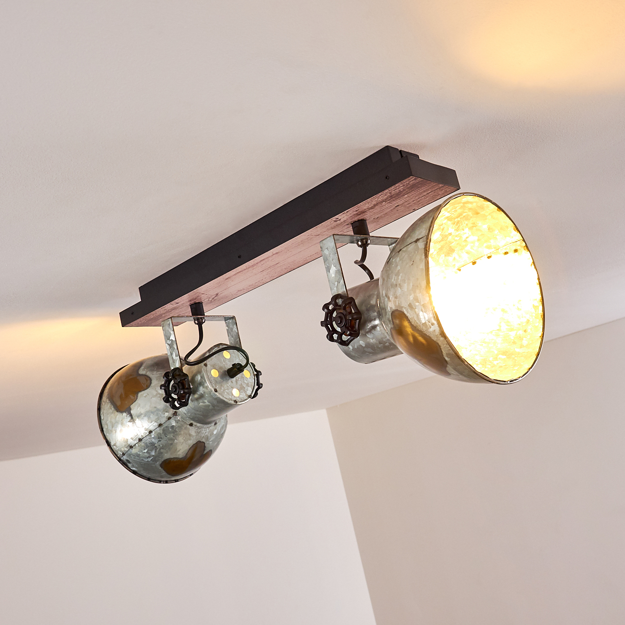 Eine Deckenleuchte mit zwei Metall-Strahlern. Die Leuchte hat einen Holzbalken mit Schwarz- und Brauntönen. Die Lichter sind an.