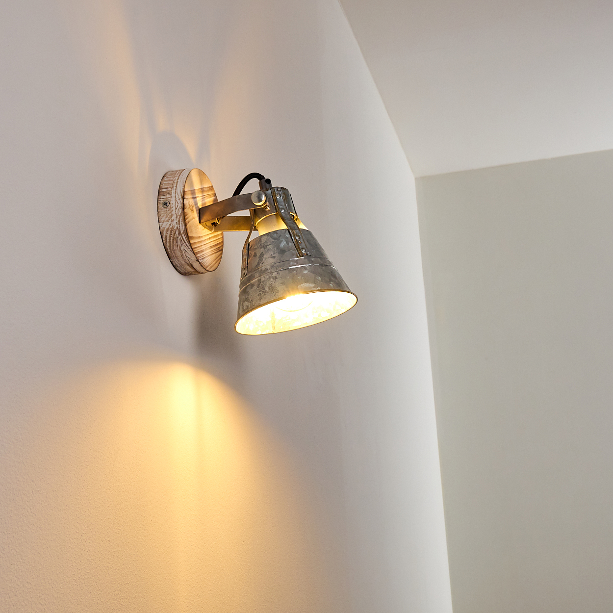 Eine Wandleuchte mit Metallschirm und Holzsockel beleuchtet eine Ecke. Das Licht wirft einen warmen Schein an die Wand.