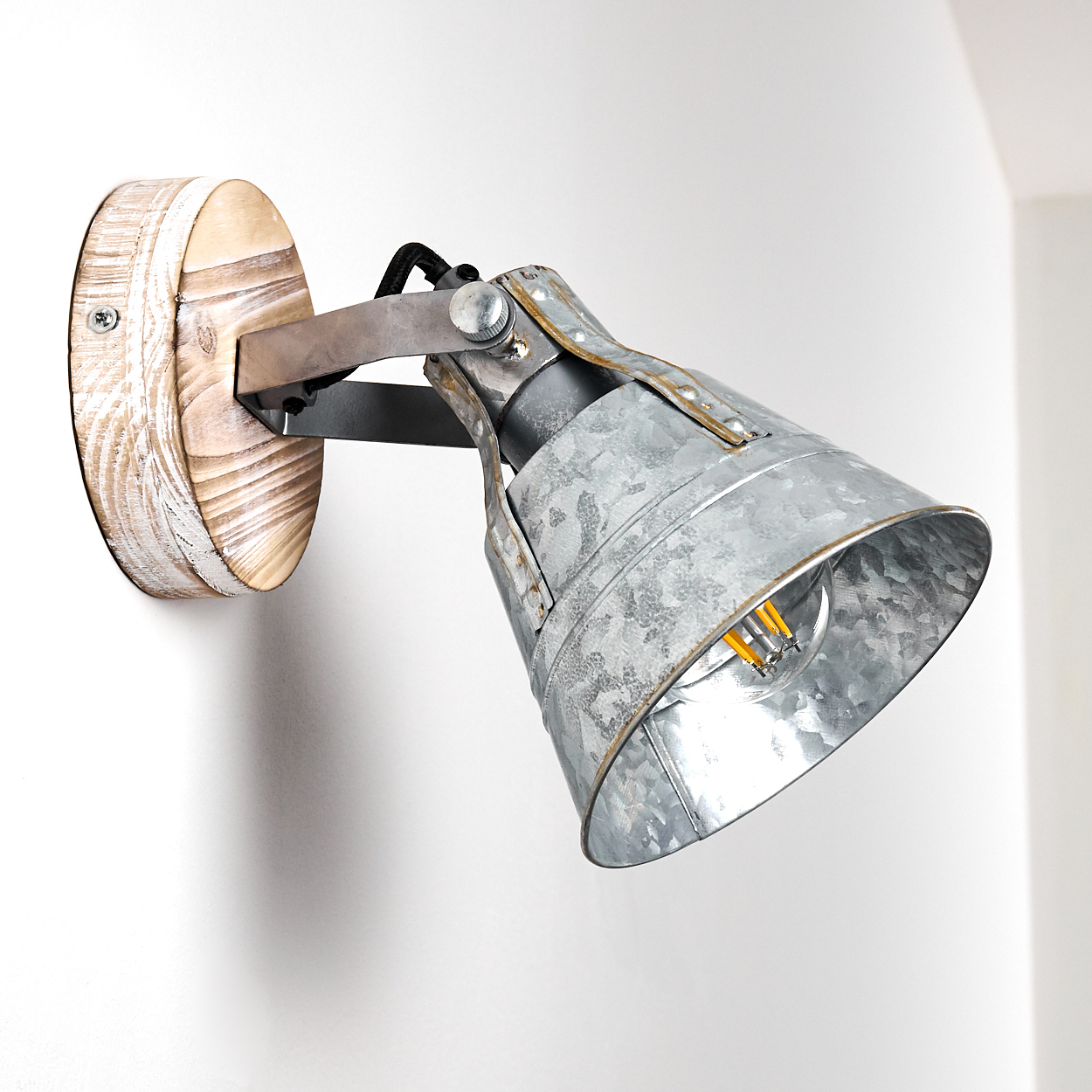 Eine Wandleuchte aus Metall und Holz. Die Lampe hat einen Lampenschirm aus verzinktem Metall und einen Holzsockel. Sie ist an einer weißen Wand montiert.