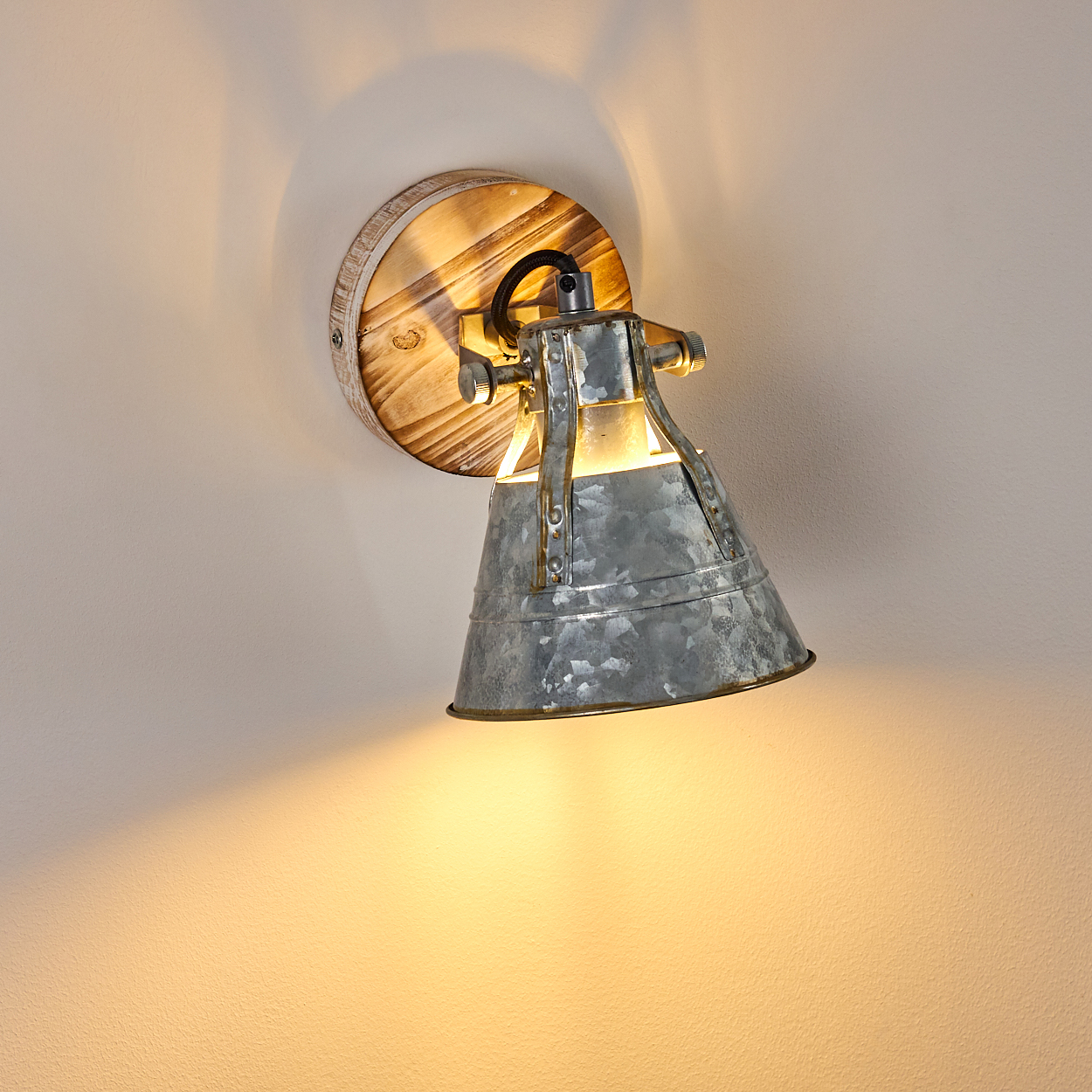 Eine Metall-Wandlampe mit Holzsockel. Die Lampe ist an und wirft ein warmes Licht auf die hellfarbene Wand.