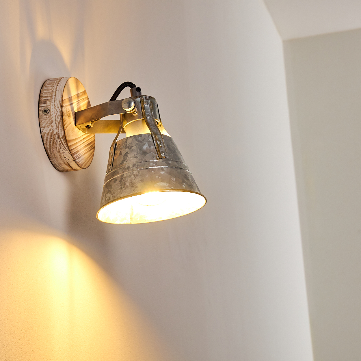 Eine Wandleuchte mit Holzsockel und Metallschirm. Die Lampe ist eingeschaltet und wirft ein warmes Licht auf die weiße Wand.