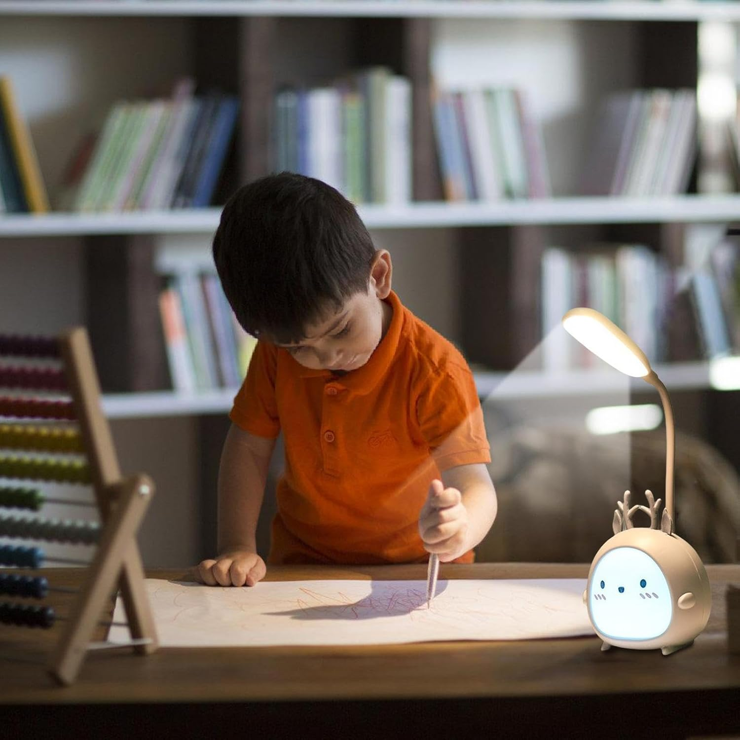 Ein Kind in orangefarbenem Hemd zeichnet auf Papier. Eine niedliche Lampe mit Hirschgeweih beleuchtet den Tisch. Bücherregale sind im Hintergrund.