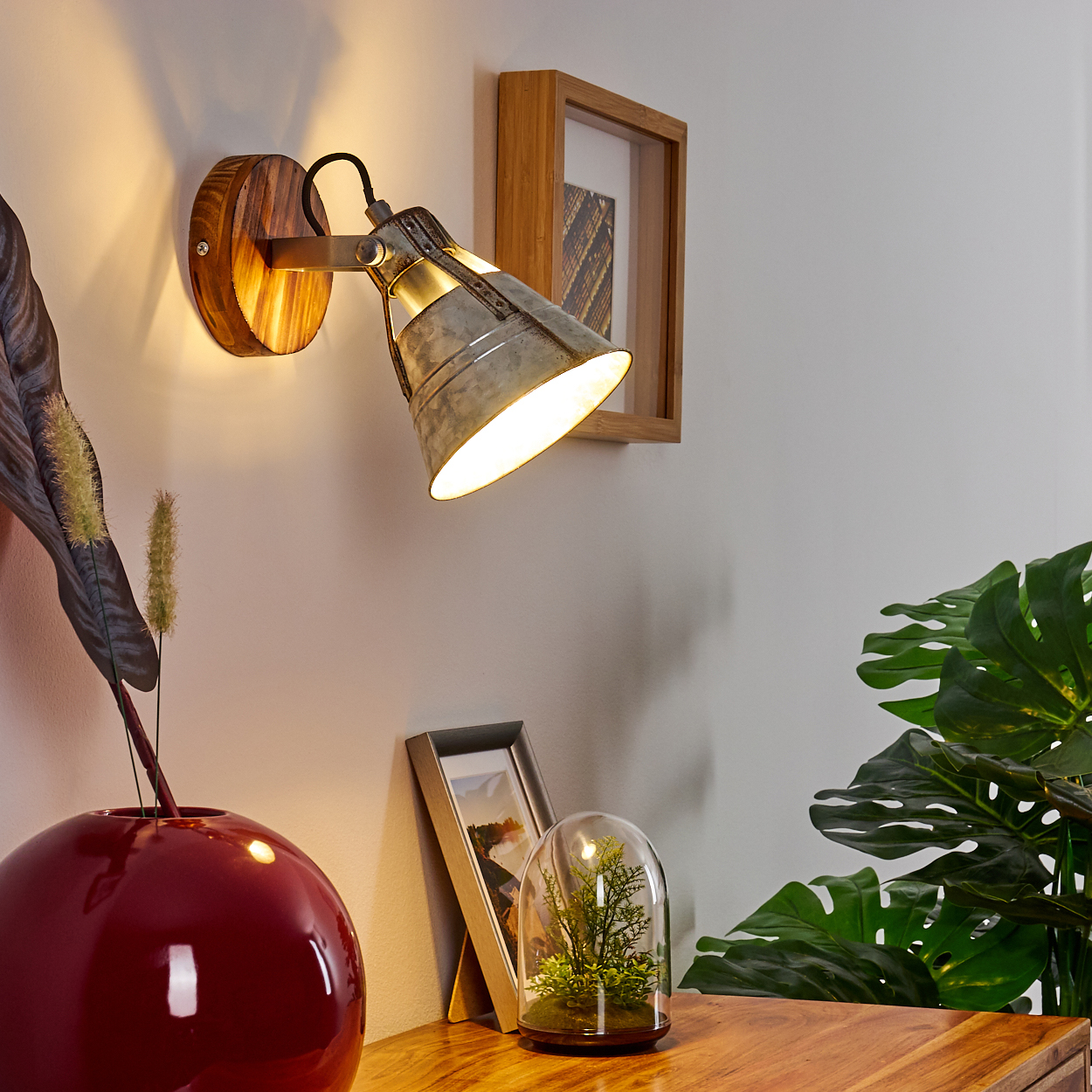 Eine Wandlampe mit Holzsockel beleuchtet einen Holztisch mit einer roten Vase, einem gerahmten Bild und einer Pflanze in einer Glaskuppel.