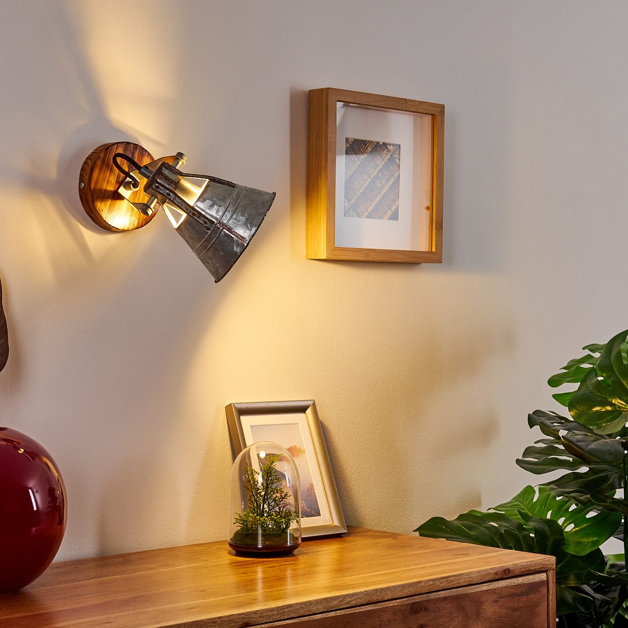 Eine Wandlampe beleuchtet eine Holzoberfläche mit Dekoration. Ein Bilderrahmen und eine Pflanze befinden sich ebenfalls auf der Holzoberfläche.