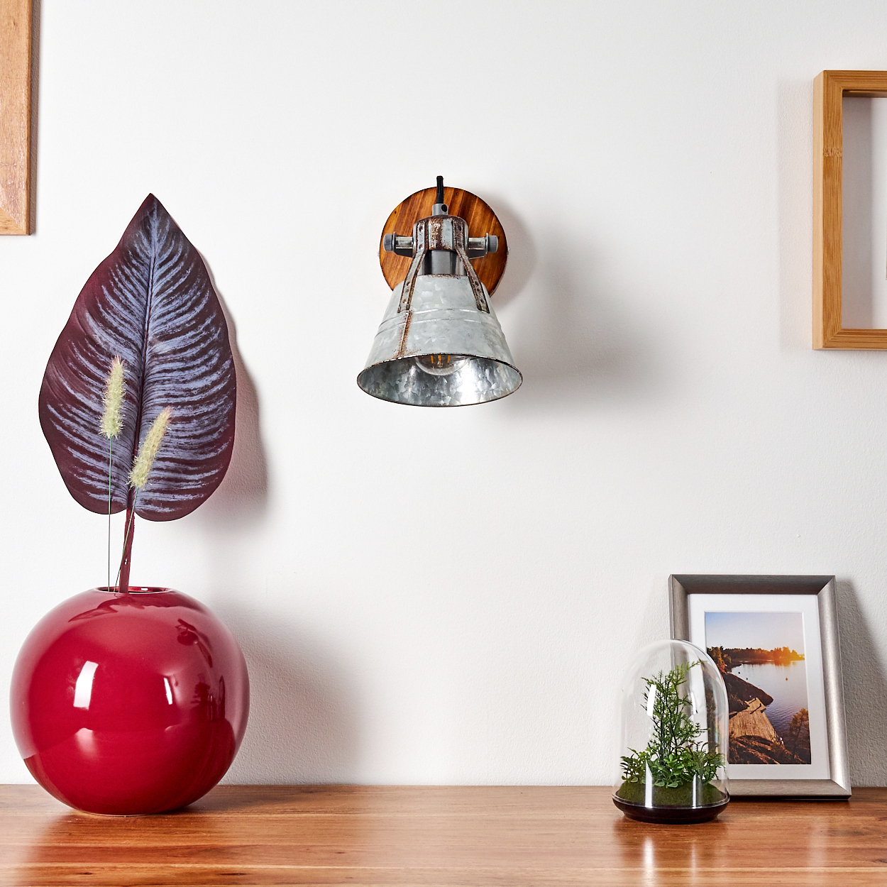 Eine rote Vase mit einem Blatt, eine Metalllampe, ein gerahmtes Bild und eine Glaskuppel mit Pflanzen auf einem Holztisch vor einer weißen Wand.