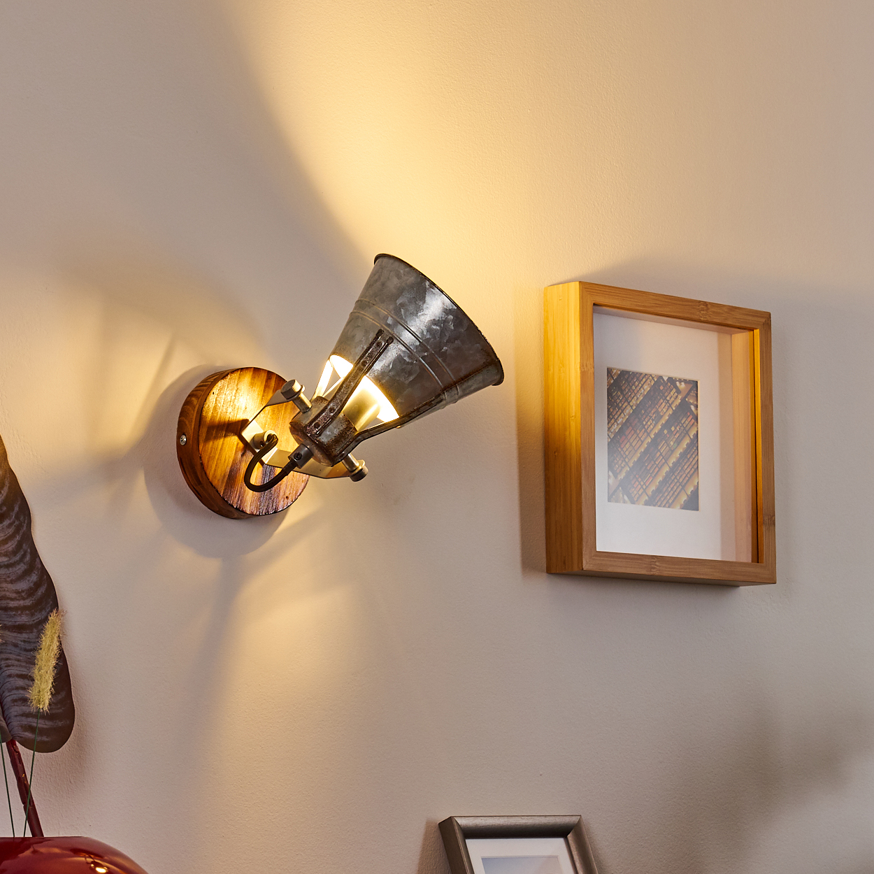 Ein an der Wand montierter Strahler mit Holzsockel und Metallschirm. Rechts hängt ein gerahmtes Bild, links ein rotes Deko-Objekt.