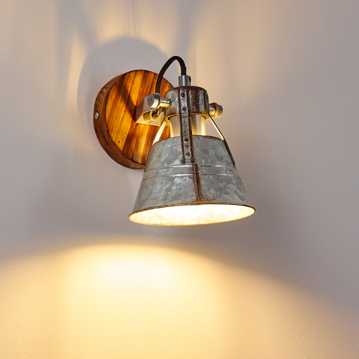 Eine rustikale Wandlampe mit Metallschirm und Holzrücken wirft ein warmes Licht auf eine weiße Wand.