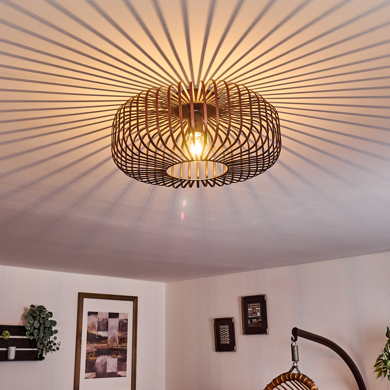 Eine moderne Deckenlampe mit kreisförmigem Design, die radiale Schatten auf die Decke wirft. Unten sind gerahmte Bilder und ein Hängestuhl zu sehen.