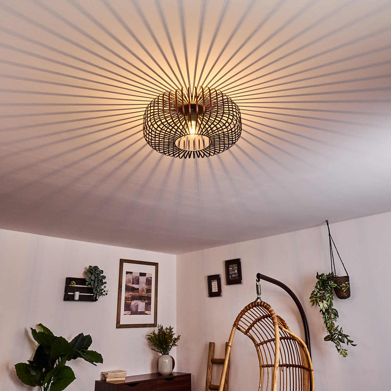 Ein modernes Zimmer mit Deckenleuchte, die Schatten wirft. Es gibt einen Rattanstuhl, Pflanzen und gerahmte Bilder an der Wand.