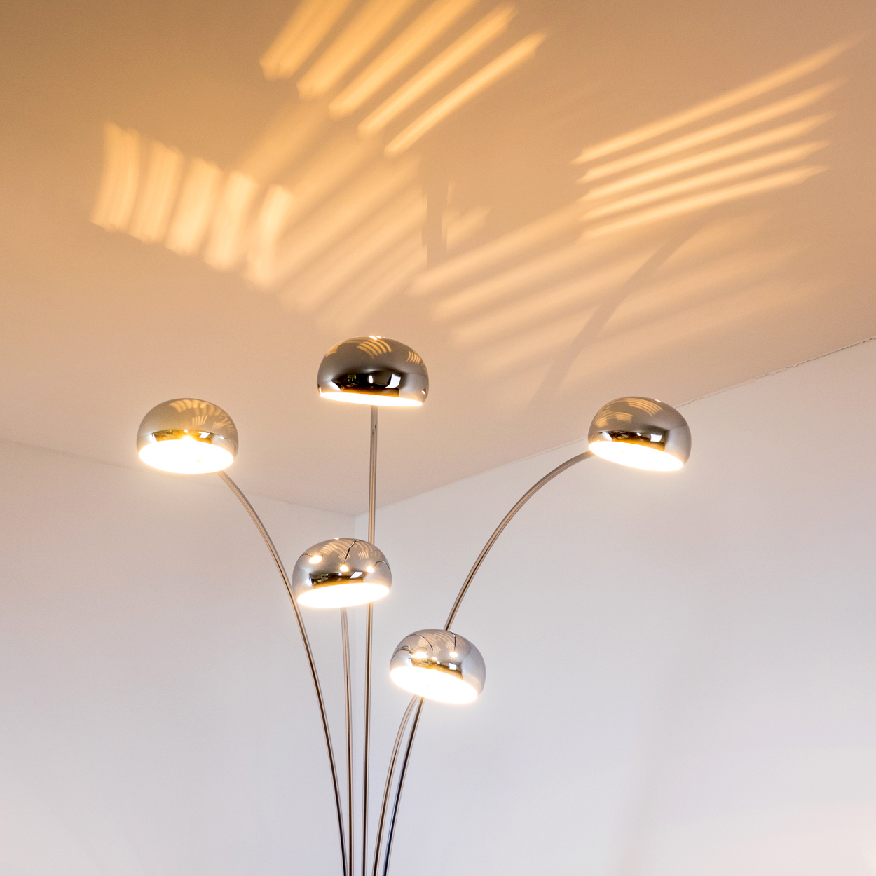 Eine moderne Stehlampe mit fünf verchromten Kuppellampen. Die Decke reflektiert das Licht, mit Schatten auf der Oberfläche.