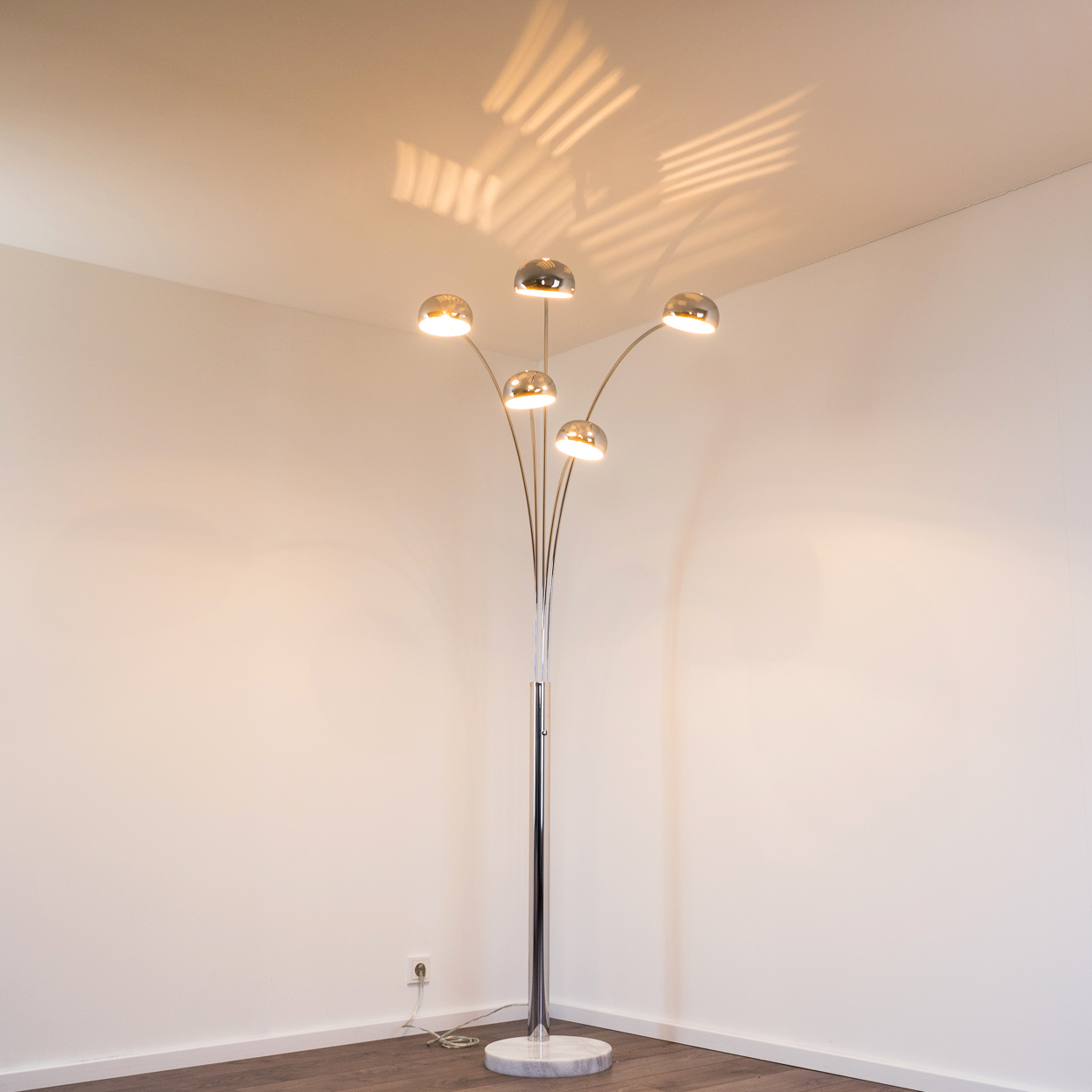 Eine verchromte Stehlampe mit fünf Lichtern. Der Sockel ist aus Marmor. Die Wand ist weiß und der Boden braun.