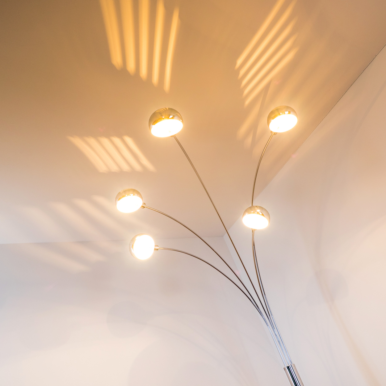 Eine moderne Stehlampe mit fünf Lichtern. Die Lampen sind kugelförmig, mit einem Chromständer und Armen. Licht wirft Schatten auf die Decke.