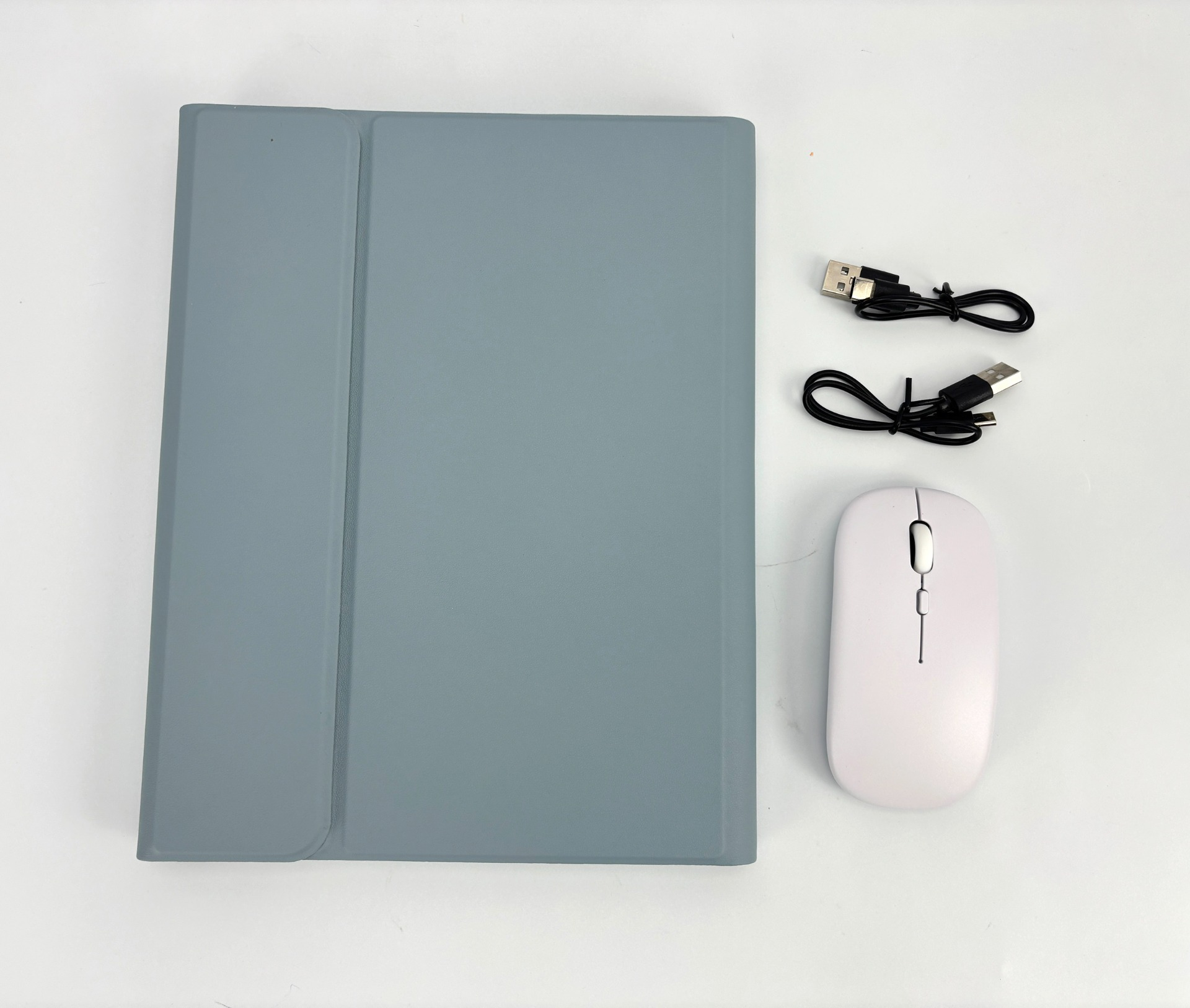 Eine Tablethülle, eine Maus und Kabel auf weißer Oberfläche. Die Hülle ist grau, die Maus ist weiß. Kabel sind schwarz.