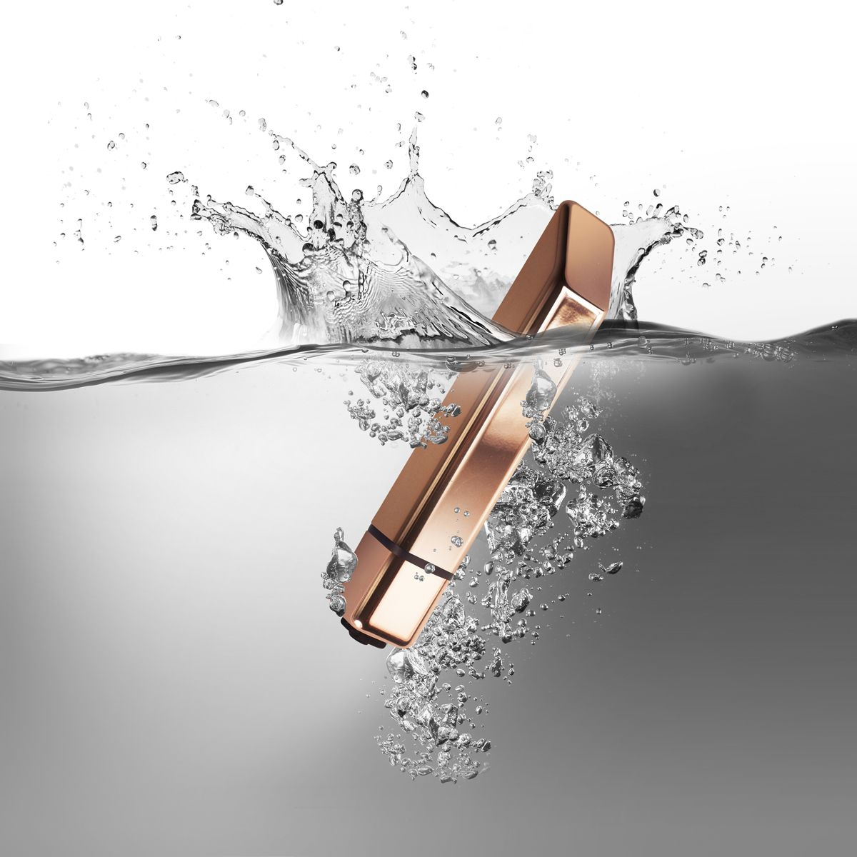 Ein roségoldenes Objekt ist teilweise in Wasser getaucht, wodurch Spritzer und Blasen entstehen. Der Hintergrund ist ein Grau-Weiß-Verlauf.