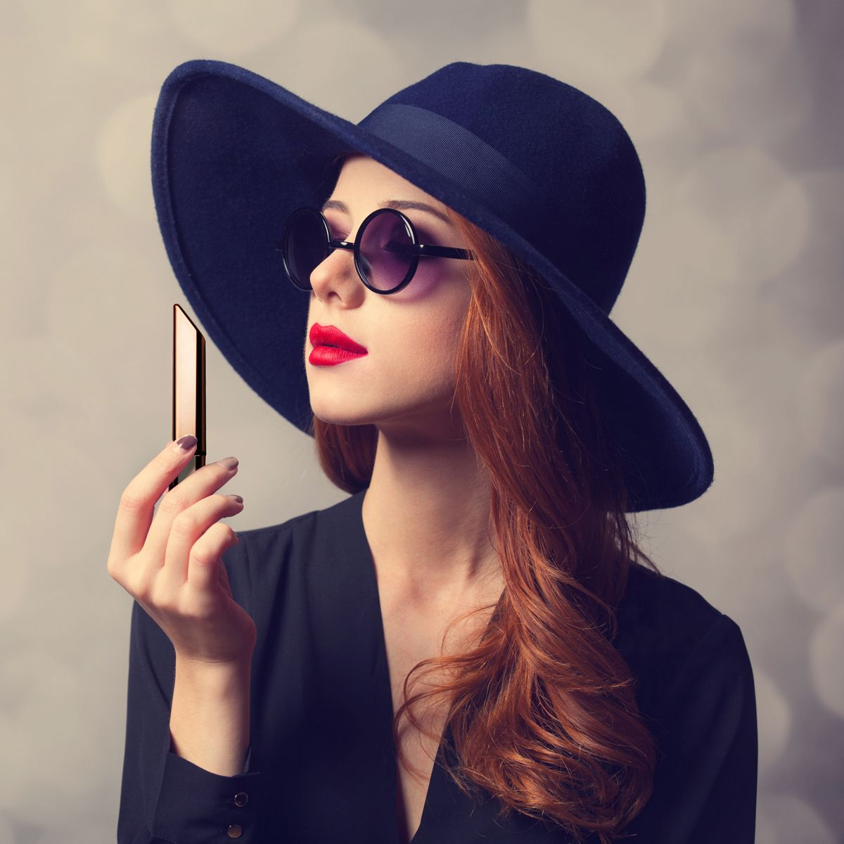 Frau mit Hut und Sonnenbrille hält Lippenstift. Sie hat rotes Haar und Lippen. Trägt ein schwarzes Oberteil, vor unscharfem Hintergrund.