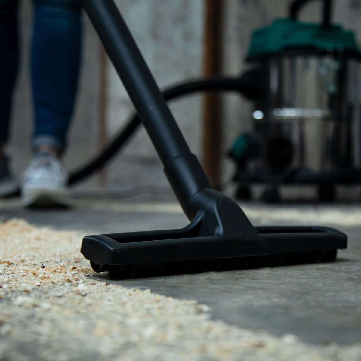 A black vacuum cleaner head on a concrete floor with wood shavings. In the background, there is a person and a vacuum.