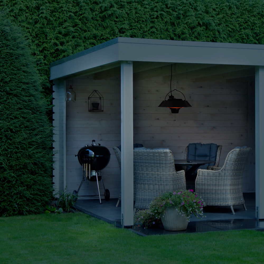 Een prieel met een grill en rotan meubels. Een hanglamp verlicht de zithoek. Groen gras en bladeren omringen de structuur.