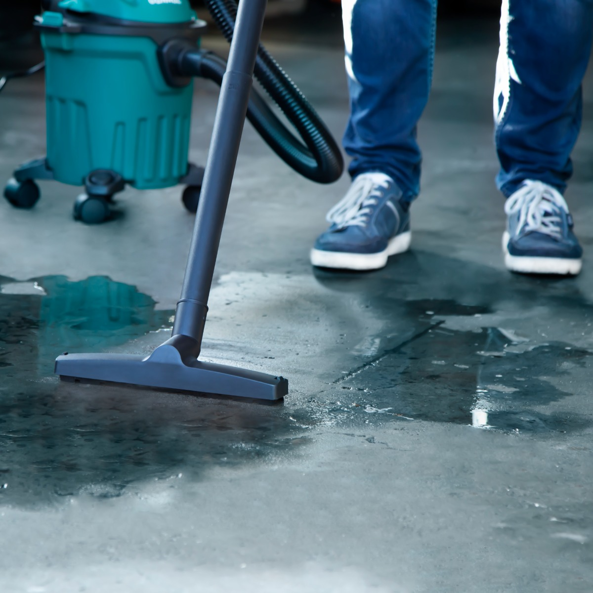 Person vacuuming a wet floor. A teal vacuum cleaner stands in the background. The person wears blue jeans and blue sneakers.