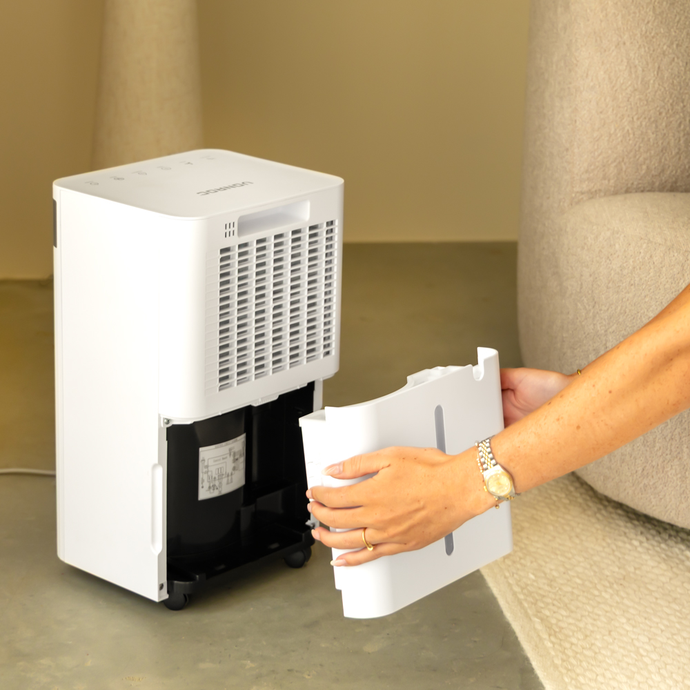 A white dehumidifier has its water tank removed by a person. The dehumidifier is on a gray floor. A beige sofa is in the background.