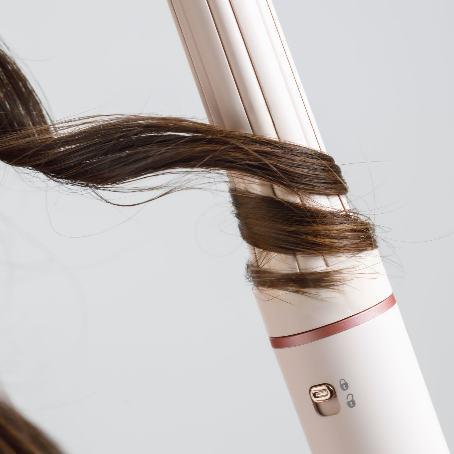 Un arricciacapelli avvolge capelli castani. Il ferro è bianco con un accento oro rosa. Lo sfondo è grigio tenue.