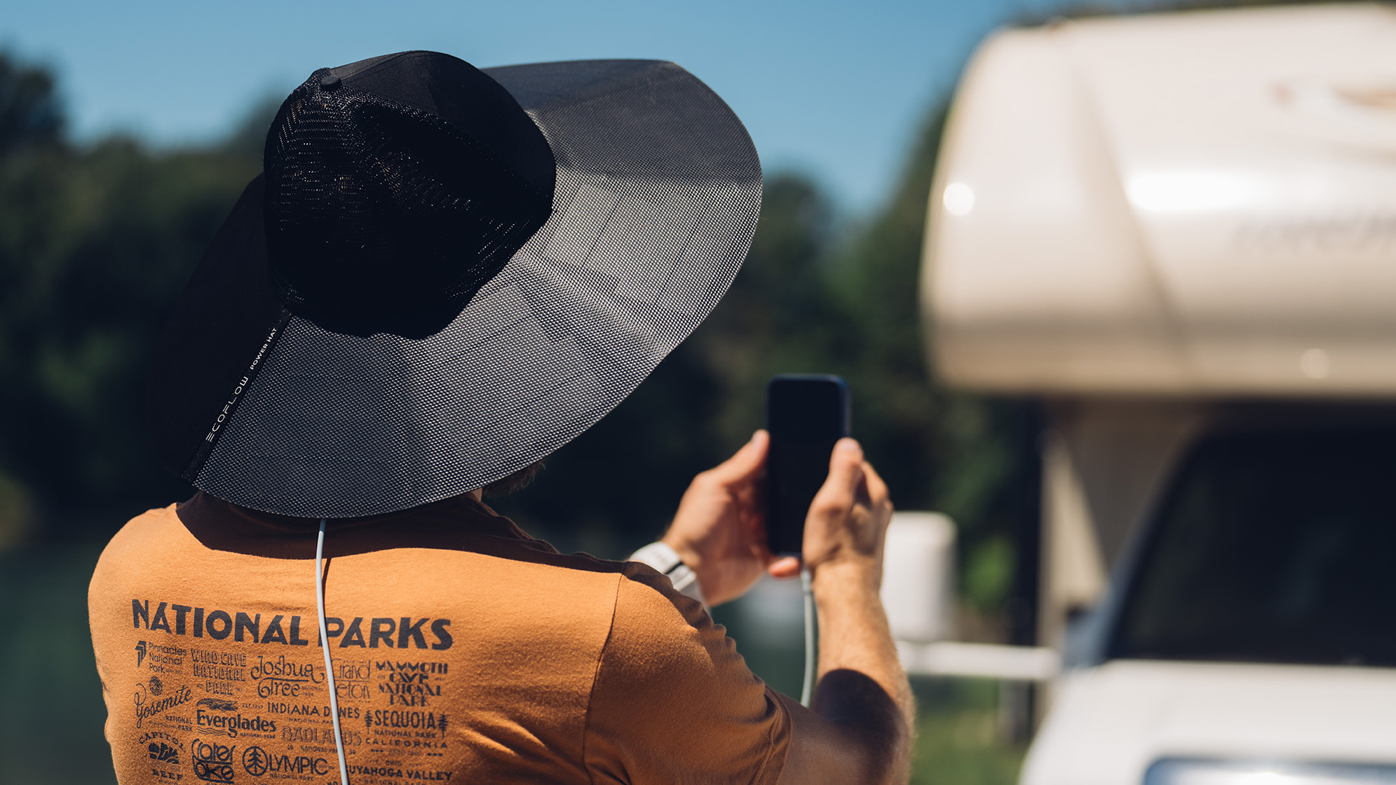 Person mit Hut, die ein Telefon benutzt und es in der Nähe eines Wohnmobils auflädt. Im Hintergrund Bäume und sonniger Himmel.