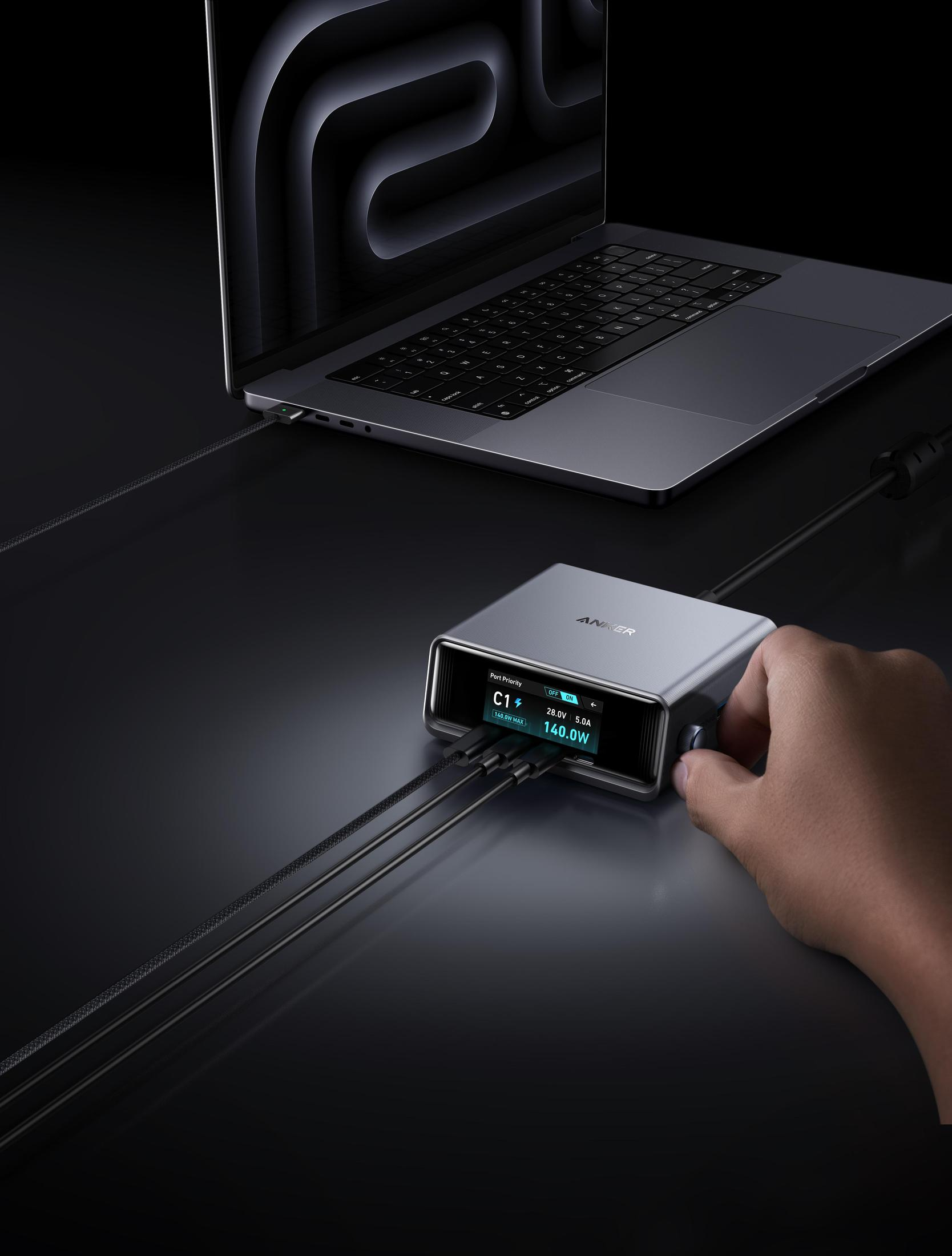 Laptop podłączony do srebrnej ładowarki Anker. Ręka reguluje ładowarkę, wyświetlacz pokazuje statystyki zasilania. Czarne kable łączą.