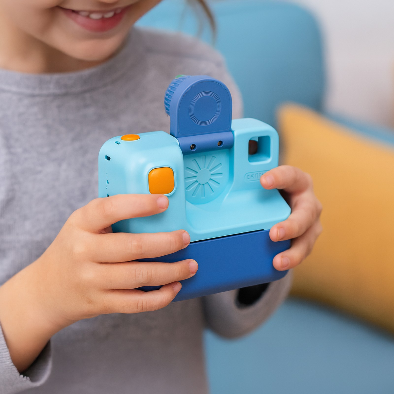 Dziecko trzyma niebiesko-pomarańczowy aparat. Dziecko się uśmiecha. W tle znajduje się niebieska kanapa.