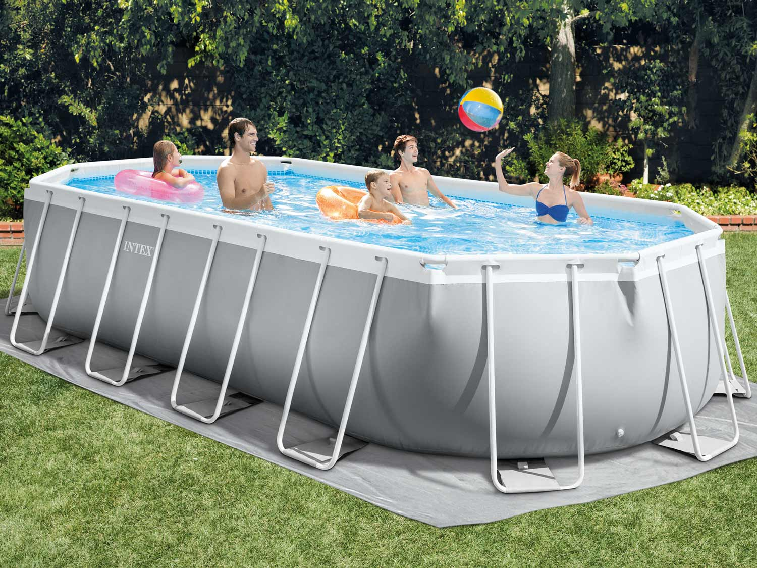 Ludzie w szarym prostokątnym basenie, bawią się piłką. Basen stoi na zielonej trawie. W tle drzewa.